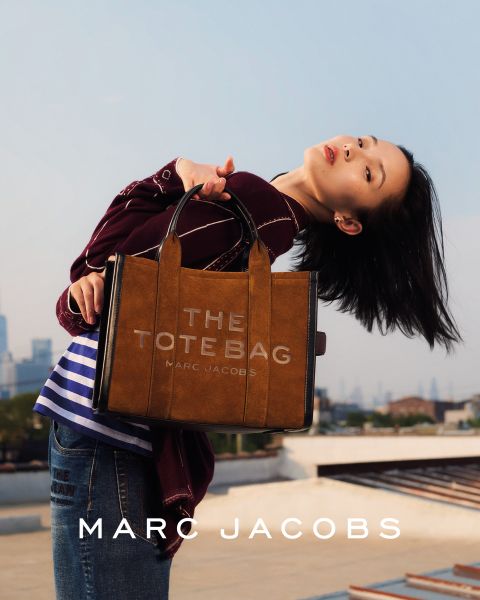 Una donna posa all'aperto con in mano una borsa marrone Marc Jacobs, si appoggia all'indietro con la testa inclinata e indossa una giacca color castagna e jeans blu. Nell'immagine compaiono le scritte "THE TOTE BAG MARC JACOBS" e "MARC JACOBS".