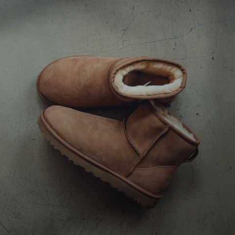 Un paio di stivali invernali in pelle scamosciata marrone chiaro con una spessa fodera in pile chiaro, appoggiati su un pavimento di cemento grigio leggermente screpolato.