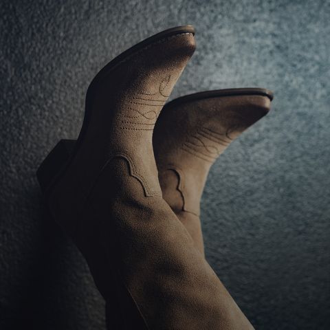 Primo piano di un paio di stivali da cowboy marroni in pelle scamosciata con cuciture decorative su uno sfondo scuro e strutturato. Gli stivali sono rivolti verso l'alto, evidenziandone il design e il materiale.