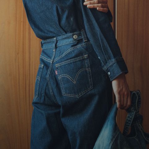 Una persona che indossa una camicia di jeans scura e jeans è in piedi di fronte a una parete di legno e tiene una giacca di jeans in una mano mentre è di spalle alla telecamera.