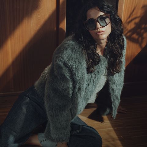 Una donna con capelli scuri ondulati che indossa occhiali da sole oversize, una giacca di pelliccia grigia e jeans scuri è seduta su un pavimento di legno vicino a una parete di legno con un'illuminazione drammatica.