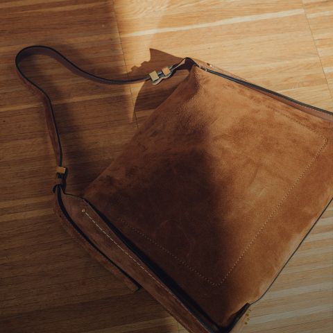 Una borsa a tracolla in pelle scamosciata marrone con un cinturino largo è appoggiata su un pavimento di legno illuminato dal sole, dove luci e ombre creano un aspetto caldo e strutturato.