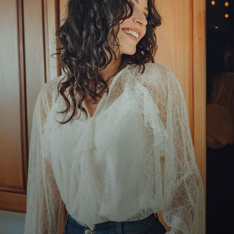 Una donna con capelli scuri ondulati, che indossa una camicetta di pizzo bianca e jeans blu, è in piedi sorridendo in una stanza con pannelli di legno illuminata in modo caldo e guarda di lato.