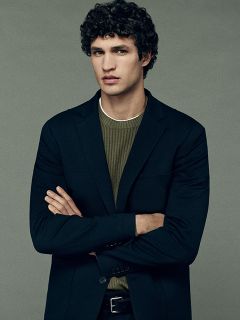 Un uomo con capelli scuri e ricci, che indossa un blazer nero sopra un maglione verde a coste, sta in piedi con le braccia conserte davanti a uno sfondo grigio uniforme e guarda dritto davanti a sé con un'espressione neutra.