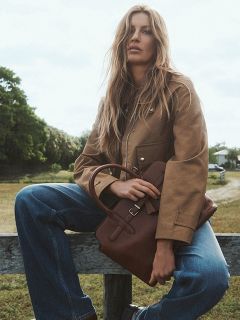 Una donna con lunghi capelli biondi è seduta all'aperto su una staccionata di legno, indossa una giacca marrone chiaro, jeans blu e tiene una borsa marrone in grembo. Sullo sfondo si vedono alberi e un sentiero.