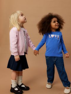 Due bambini piccoli stanno in piedi tenendosi per mano davanti a uno sfondo marrone uniforme. Un bambino indossa una giacca rosa e una gonna, l'altro jeans blu e una camicia blu con la scritta "L'AMORE È BAMBINO". Entrambi guardano in su con curiosità.
