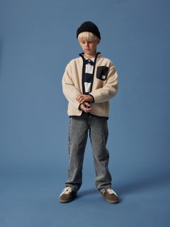 Un ragazzo con i capelli biondi indossa un berretto nero, un pile color crema sopra una camicia a righe, jeans grigi e scarpe da ginnastica. È in piedi di fronte a uno sfondo blu uniforme e guarda in basso con le mani giunte.