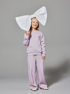 Una giovane ragazza sorridente in tuta color lavanda posa su uno sfondo grigio con un grande fiocco bianco in testa.