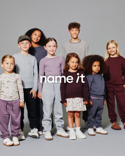Un gruppo di otto bambini diversi è in piedi insieme su uno sfondo grigio, tutti indossano abiti casual coordinati in colori neutri e pastello. Il testo "name it" appare al centro dell'immagine.