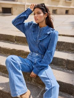 Donna seduta su una scalinata di pietra all'aperto, indossa una camicetta di jeans a maniche lunghe con dettagli a volant e jeans a vita alta abbinati. Ha i capelli scuri, occhiali da sole sulla testa e guarda pensierosa in lontananza.
