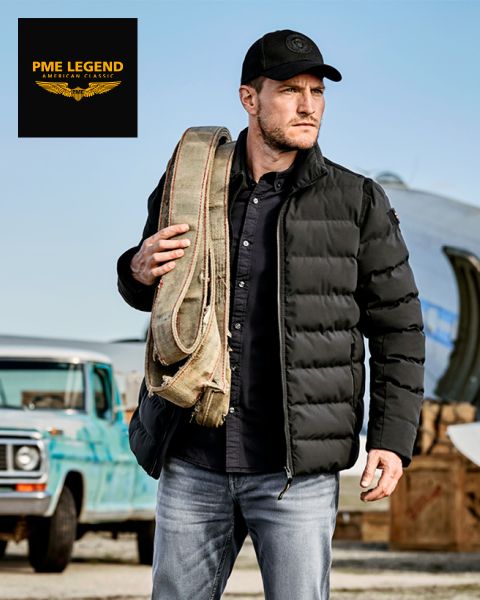 Un uomo con un piumino nero, un cappello e dei jeans porta una cinghia di tela arrotolata sulla spalla e si trova all'aperto vicino a un vecchio camion blu e un aereo. Il logo di PME Legend appare nell'angolo in alto a sinistra.