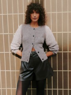 Una donna con i capelli ricci è in piedi accanto a una parete piastrellata. Indossa un cardigan grigio chiaro e soffice, pantaloncini di pelle neri a vita alta e collant neri, con le mani in tasca e un'espressione sicura di sé.