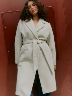 Una persona con i capelli ricci e una cintura beige chiaro è in piedi davanti a un muro rosso-marrone e guarda leggermente verso il basso con un'espressione rilassata.