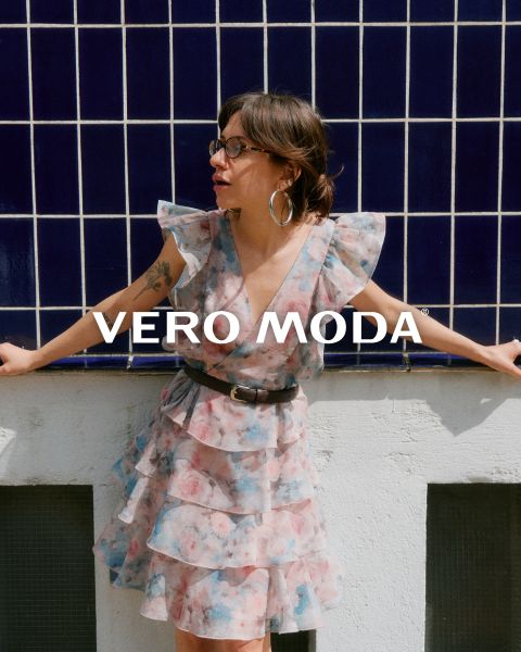 Una donna con gli occhiali e un abito floreale con volant è appoggiata a un muro bianco davanti a uno sfondo piastrellato di blu. La scritta "VERO MODA" appare al centro dell'immagine.