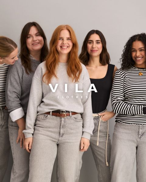 Cinque donne in abbigliamento casual, tra cui jeans e top in colori neutri, sono in piedi vicine e sorridono davanti a uno sfondo semplice. Il testo sovrapposto recita "Vila Clothes".