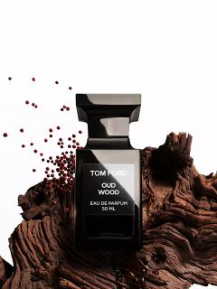 Un flacone nero del profumo Tom Ford Oud Wood è posizionato su un pezzo di legno strutturato, vicino al quale sono sparsi piccoli semi marroni, su uno sfondo bianco.