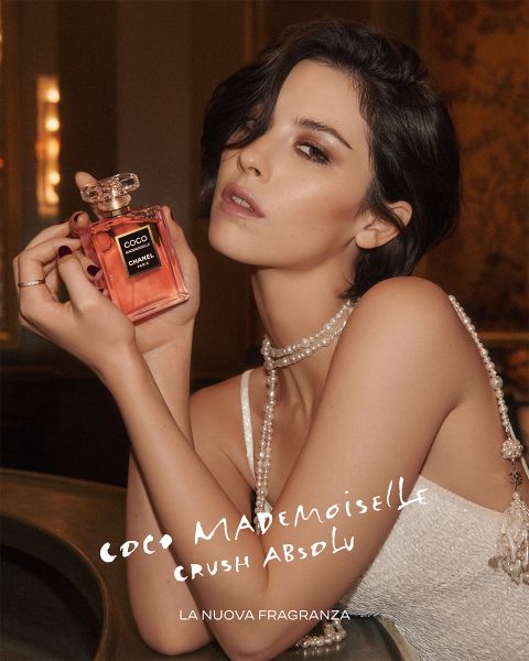 Una donna in un abito elegante tiene in mano una bottiglia del profumo "Coco Mademoiselle" di Chanel e guarda la telecamera. Il testo pubblicizza la fragranza in italiano.