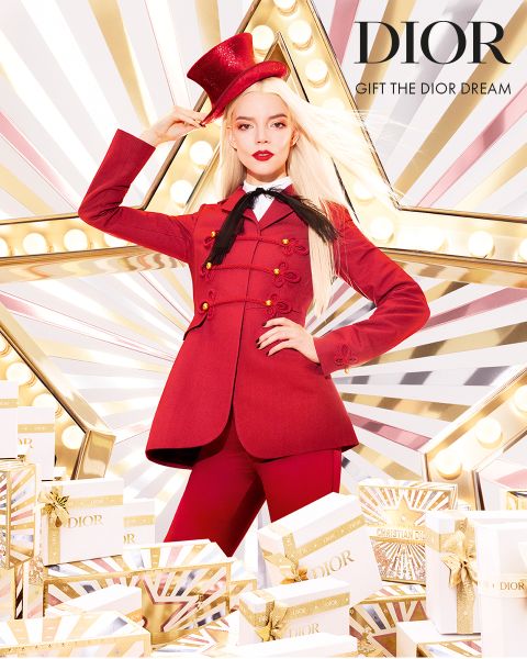 Una donna in un vistoso abito rosso e cilindro posa con sicurezza davanti a una grande stella illuminata, circondata da confezioni regalo Dior bianche e dorate. Il testo recita "DIOR REGALA IL SOGNO DI DIOR".