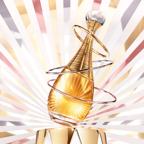 Un flacone di profumo dorato con eleganti anelli a spirale si erge di fronte a uno sfondo radioso di raggi dorati, rosa e bianchi, creando un aspetto lussuoso e glamour.