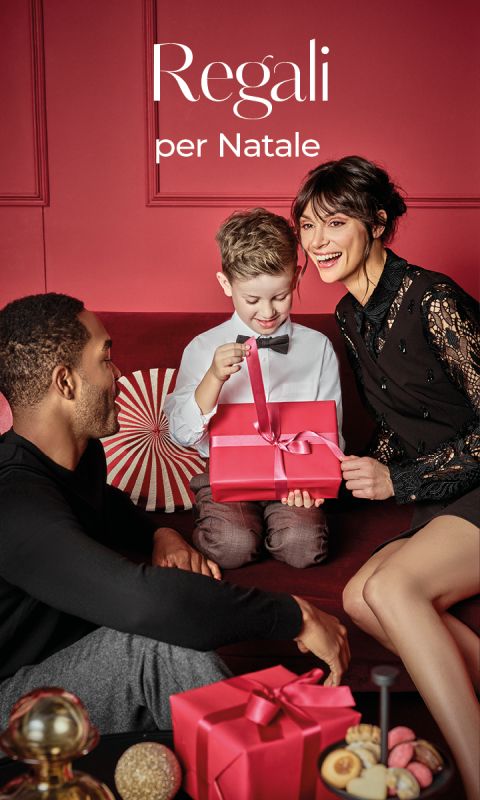 Una famiglia felice è seduta su un divano rosso e un ragazzino apre una scatola regalo rossa, circondato da decorazioni festive. Il testo di sfondo in italiano recita: "Regali per Natale".