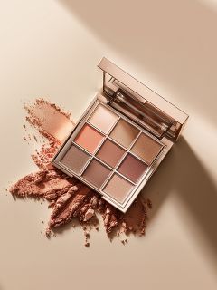 Una palette di trucco quadrata con nove ombretti in tonalità neutre è aperta su una superficie beige, dove alcune delle tonalità in polvere sono sparse e schiacciate. Nello sportello della palette si vede uno specchio.