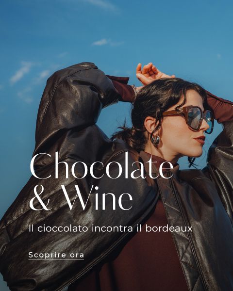 Una donna con occhiali da sole e giacca di pelle marrone posa con le mani sopra la testa davanti a un cielo blu. Il testo sovrapposto recita: "Schokolade & Wein. Il cioccolato incontra il bordeaux. Scoprire ora."