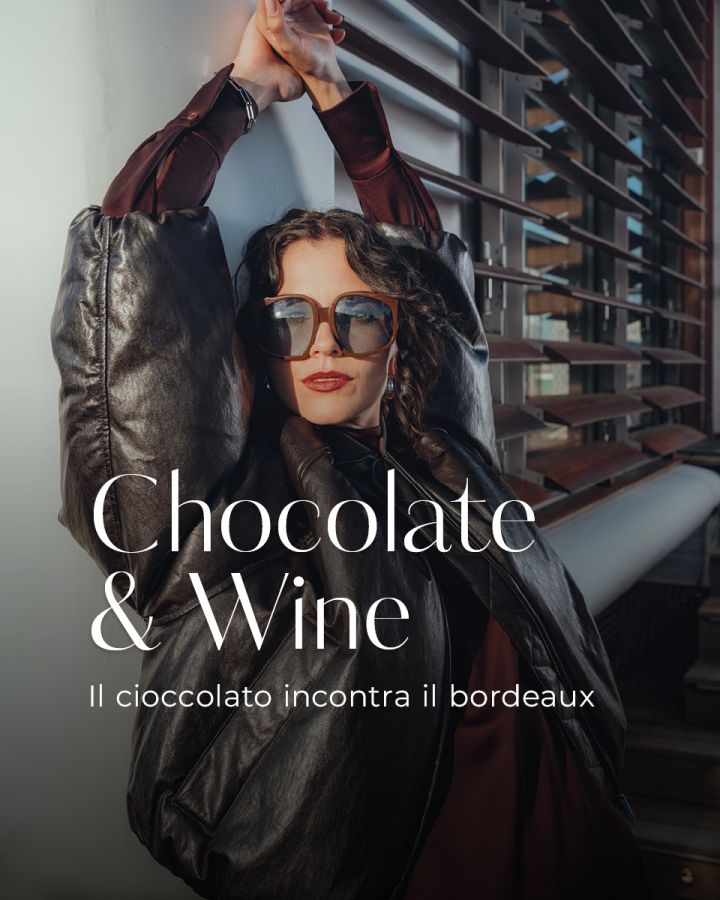 Una donna elegante con occhiali da sole e una giacca scura e voluminosa si appoggia a una ringhiera, dietro di lei linee architettoniche moderne. Le sovrapposizioni di testo recitano "Chocolate & Wine" e "Il cioccolato incontra il bordeaux".