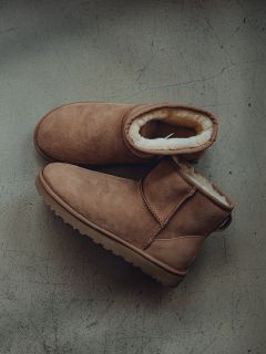 Un paio di stivali marroni in pelle scamosciata foderati in shearling sono appoggiati su un pavimento grigio strutturato, con uno stivale che si sovrappone leggermente all'altro.