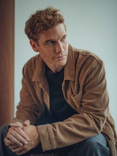 Un uomo con capelli castano chiaro ricci è seduto in una casa e indossa una giacca di velluto a coste marrone sopra una camicia nera. Guarda pensieroso di lato e ha le mani leggermente incrociate. Lo sfondo è illuminato delicatamente e neutro.