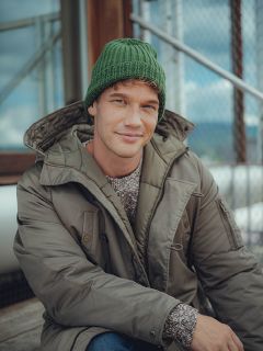 Un uomo che indossa un berretto di lana verde, un maglione grigio e una giacca invernale verde oliva è seduto all'aperto su gradini di legno e sorride dolcemente alla fotocamera. Sullo sfondo si vedono una recinzione metallica e un edificio in vetro.