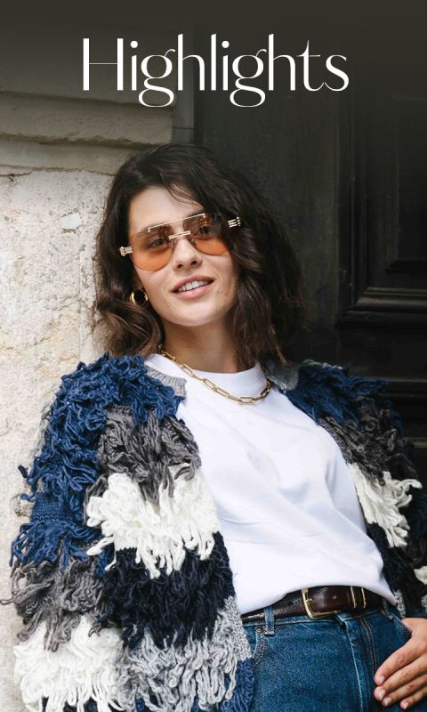 Una donna con capelli castani ondulati che indossa occhiali da sole, un grosso maglione a trecce blu e bianco, una maglietta bianca, jeans e una collana d'oro, sorride in piedi accanto a un edificio. Sopra di lei appare la parola "Highlights".