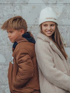 Un ragazzo e una ragazza sono schiena contro schiena davanti a un muro di pietra chiaro. Il ragazzo indossa una giacca marrone e guarda di lato, mentre la ragazza indossa un cappotto soffice e un cappello bianco e sorride mentre guarda lontano dal ragazzo.