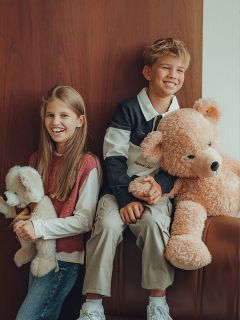 Due bambini sorridenti siedono uno accanto all'altro, ognuno con un peluche. La bambina ha i capelli lunghi e abbraccia un orsacchiotto più piccolo, mentre il bambino tiene in mano un orsacchiotto grande. Sono seduti accanto a una parete di legno e sembrano felici e rilassati.