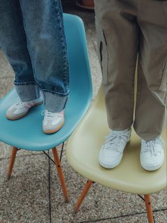 Due bambini sono in piedi su delle sedie; uno su una sedia blu, indossa jeans e scarpe da ginnastica con strisce rosa, l'altro su una sedia gialla, indossa pantaloni color kaki e scarpe da ginnastica bianche. Si vedono solo le loro gambe e i loro piedi.