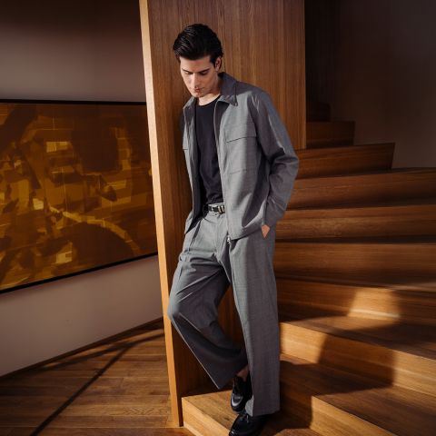 Un uomo in abito grigio con camicia nera e scarpe nere è in piedi accanto a una scala di legno e si appoggia con disinvoltura a una parete in un interno moderno.