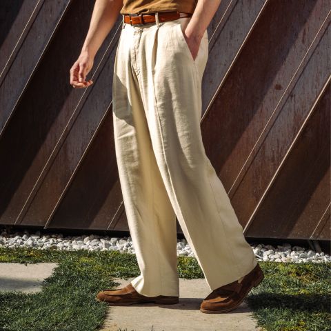 Una persona è in piedi all'aperto e indossa pantaloni a pieghe color crema, una cintura marrone, una camicia marrone chiaro e scarpe marroni, con la luce del sole che proietta ombre sul terreno.