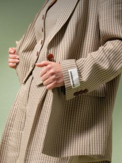 Una persona che indossa un abito a quadri beige-marrone con un'etichetta bianca con la scritta "ABANDONABLE" sul polsino, tiene il risvolto della giacca davanti a uno sfondo verde.