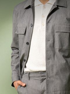 Una persona che indossa una giacca grigio chiaro abbottonata e pantaloni abbinati sopra una polo bianca testurizzata, in piedi con una mano in tasca davanti a uno sfondo verde tenue.