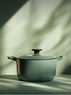 Un forno olandese in ghisa smaltata verde con coperchio e pomello nero è posizionato su una superficie illuminata da una morbida luce naturale con ombre sfocate sullo sfondo.