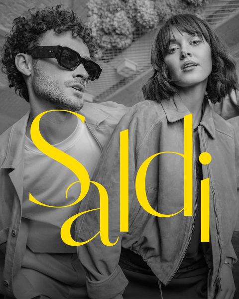 Un uomo elegante e una donna elegante posano insieme in abiti alla moda; un grande testo giallo con la scritta "Saldi" si trova sopra l'immagine in bianco e nero.