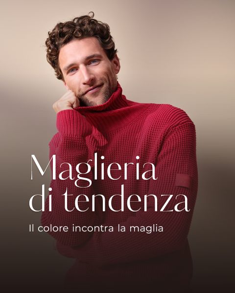 Un uomo con capelli ricci e barba indossa un dolcevita rosso acceso e sorride dolcemente con una mano sulla guancia. Maglieria di tendenza. Il colore incontra la maglia.