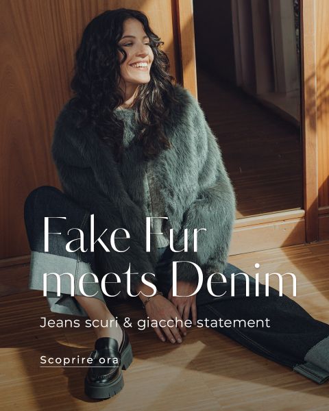 Una donna sorridente è seduta sul pavimento accanto a una porta di legno e indossa una giacca grigia in ecopelliccia, jeans scuri e stivali con plateau. Il testo sovrapposto recita: "Ecopelliccia incontra il denim. Jeans scuri & giacche statement. Scopri ora.".