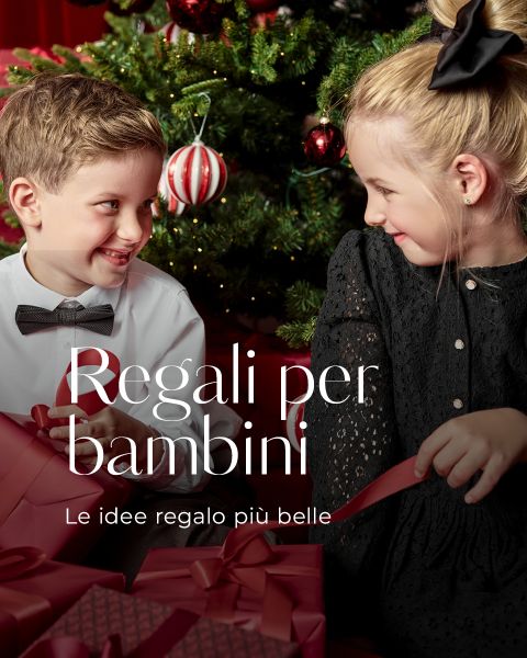 Un ragazzo in camicia bianca e papillon sorride a una ragazza in abito nero mentre sono seduti davanti a un albero di Natale decorato e tengono in mano scatole regalo rosse. Il testo italiano recita "Regali per bambini. Le idee regalo più belle.