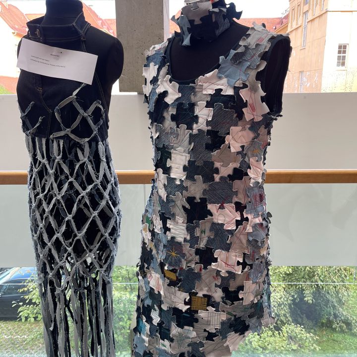 Due manichini espongono abiti creativi; uno indossa un abito a rete con fili pendenti, l'altro un abito senza maniche fatto di pezzi di puzzle colorati sovrapposti. Entrambi sono esposti davanti a uno sfondo di una finestra.