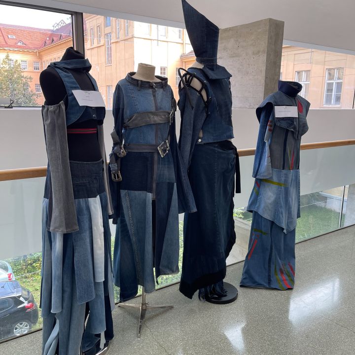 Quattro manichini espongono abiti in denim d'avanguardia con forme geometriche e dettagli multistrato, tra cui cinture larghe, cinghie e un drammatico cappello a punta. Si trovano in interni di fronte a grandi finestre con vista su edifici storici.