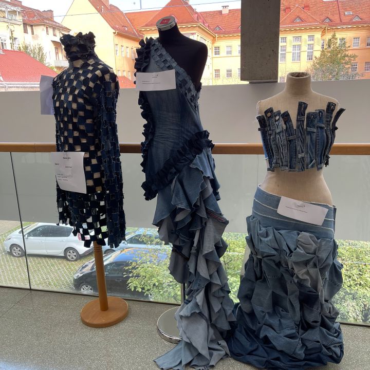 Tre manichini espongono capi creativi realizzati con denim riciclato, caratterizzati da motivi intrecciati, arricciati e multistrato. Si trovano all'interno, di fronte a grandi finestre con una vista sulla città sullo sfondo.