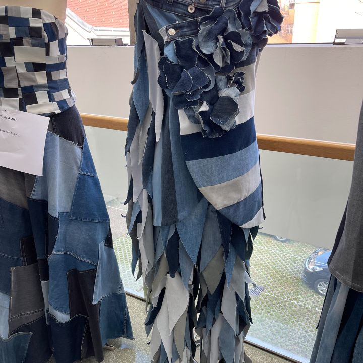 Un abito di jeans in varie tonalità esposto su un manichino, disposto a strati a forma di foglia e decorato con fiori di jeans in vita. Accanto, un altro abito patchwork di denim è parzialmente visibile.