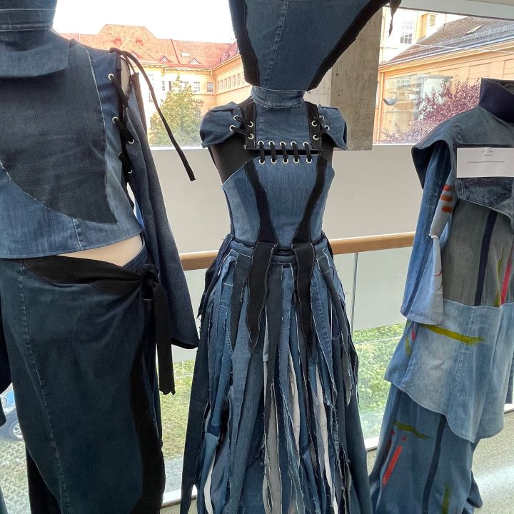 Un abito in diverse tonalità di denim con corpetto allacciato, spalle appuntite e gonna a strisce lunghe di tessuto, esposto su un manichino in una stanza accanto ad altri capi di abbigliamento in denim.