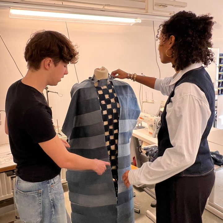 Due persone in un atelier di moda discutono un capo d'abbigliamento su un manichino. Uno indica il tessuto, che presenta strisce blu e un motivo a quadri, mentre l'altro sistema il capo. Sullo sfondo si vedono macchine da cucire e strumenti.