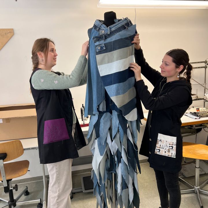 Due donne sistemano un capo di abbigliamento in jeans con motivi a righe e a zig zag su un manichino in un laboratorio, circondate da sedie e macchine da cucire.